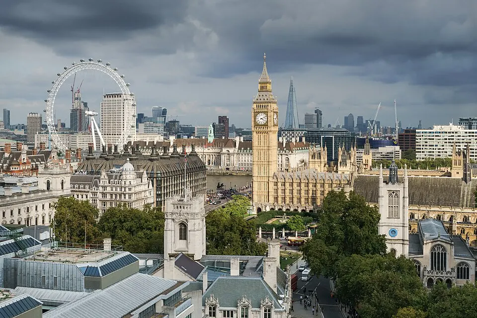 A Gothic stone parliament building with a tall clock tower, seen over rooftops with a big wheel in the distance.