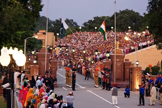 Soldiers in dress uniform at an open gate while crowds fill the stands on both sides of a road at dusk.