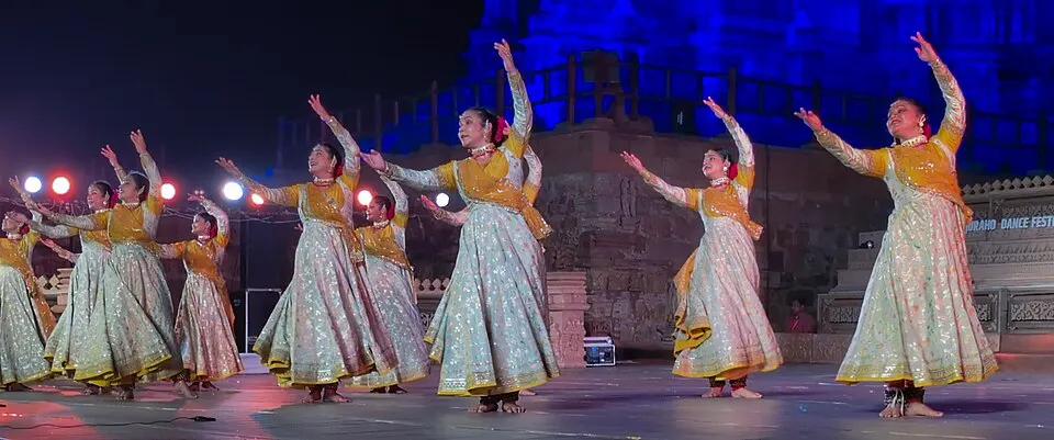 मंच पर सुनहरे-रुपहले परिधानों में कई नृत्यांगनाएँ एक साथ हाथ उठाकर नृत्य करती हुई।