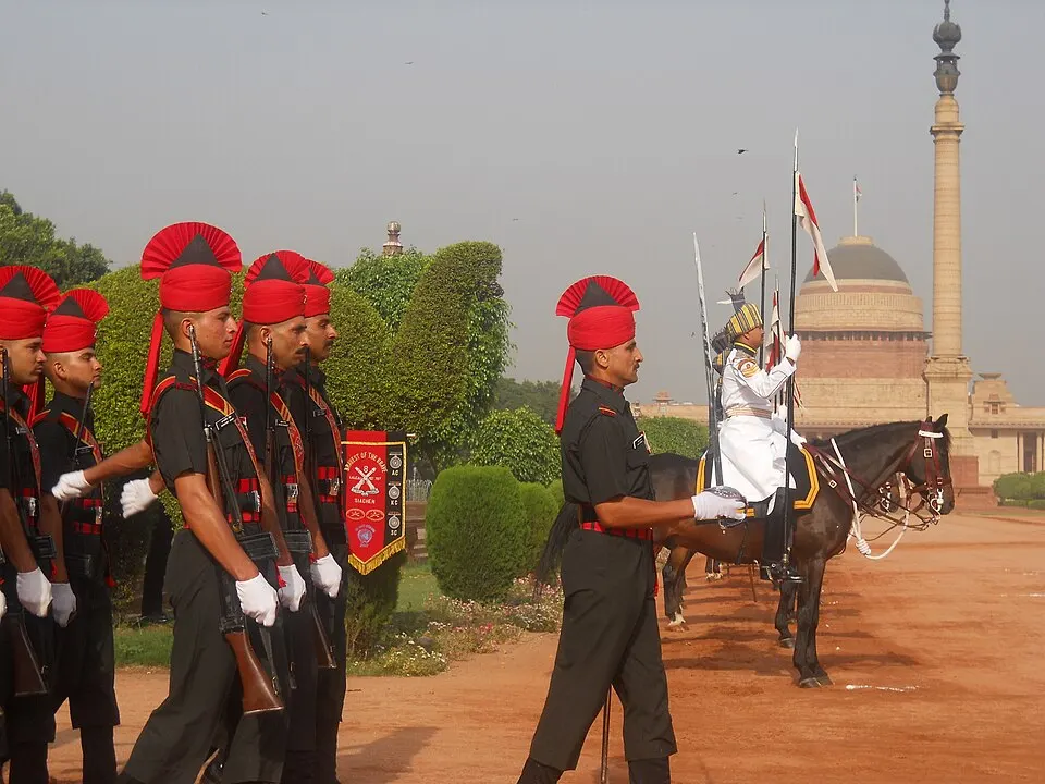 Soldiers in black uniforms and red turbans march in line near a mounted horseman in white, with a domed sandstone building and tall pillar behind.