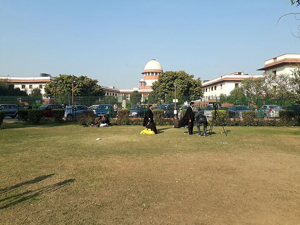 A white-domed court building behind a green fence, with lawyers in black gowns on a lawn in front.