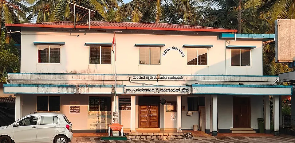 A two-storey white village office building with a red roof, coconut palms behind and a car parked in front.