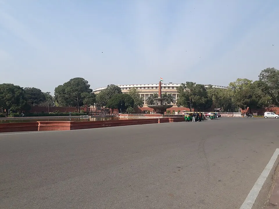 A distant round colonnaded building with a flag, seen across a wide road, a fountain and trees.