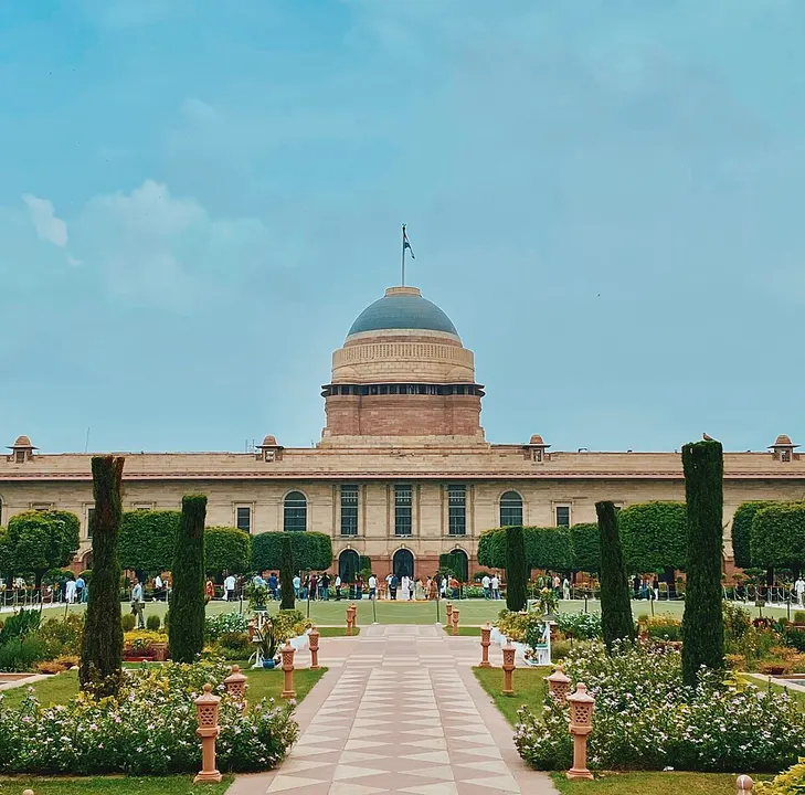 A grand sandstone palace with a large grey dome seen across a paved path and formal gardens.