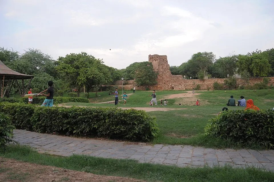 Children playing a game of cricket on a grassy park with old stone ruins in the background.