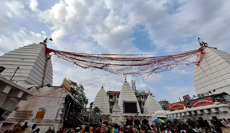 Tall white tiered temple spires rise above a crowd of pilgrims, with red threads strung overhead.
