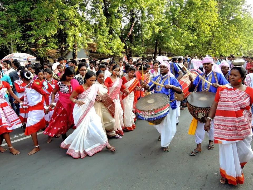 Women in white saris with red borders dance beside drummers in blue under leafy green trees on a road.