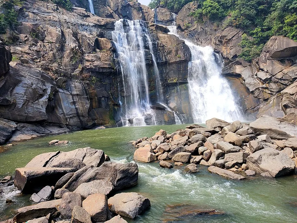 Two streams of water drop over dark rock cliffs into a green pool strewn with large boulders.