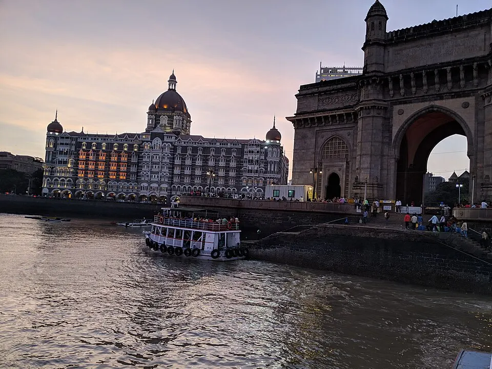 शाम के समय समुद्र के किनारे पत्थर का विशाल मेहराबदार द्वार, पास में एक भव्य होटल की इमारत और पानी में नाव।