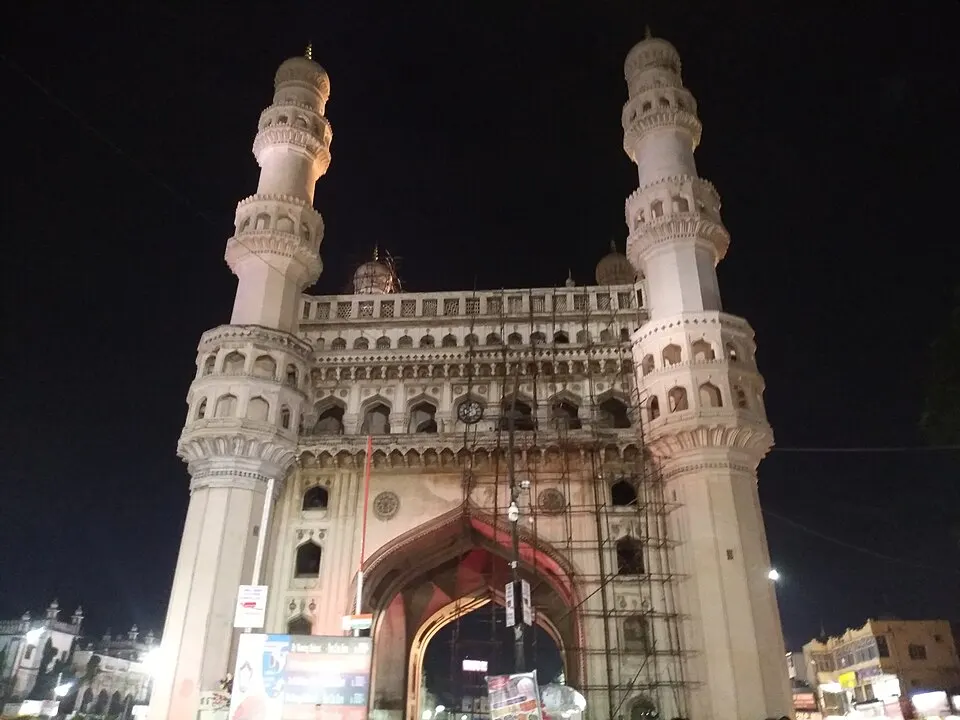 रात की रोशनी में चार ऊँची मीनारों और बड़े मेहराब वाली सफ़ेद ऐतिहासिक इमारत, नीचे बाज़ार की हलचल।