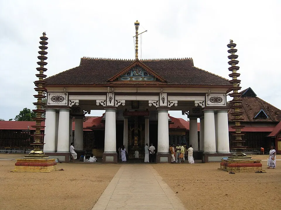 A temple entrance with white pillars and a tiled sloping roof stands between two tall tiered brass lamp towers on a sandy courtyard.