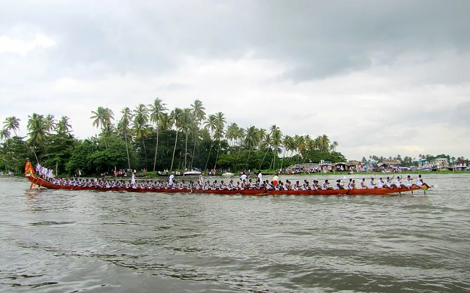 A very long red snake boat packed with rowers in white glides across a backwater lined with coconut palms.