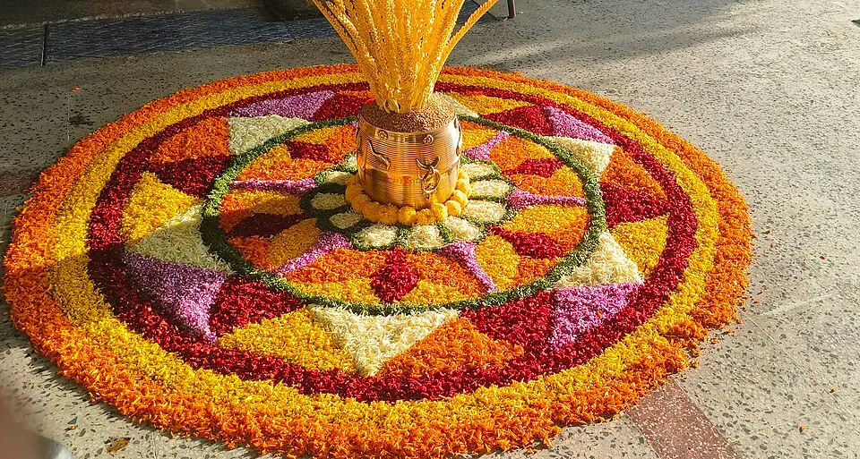 A big circular carpet of orange, yellow, red and white flower petals surrounds a brass pot filled with a sheaf of paddy.