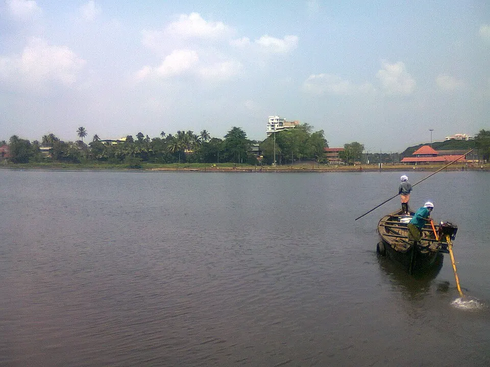 Two men in a small wooden boat on a broad, calm river with a palm-lined bank in the distance.
