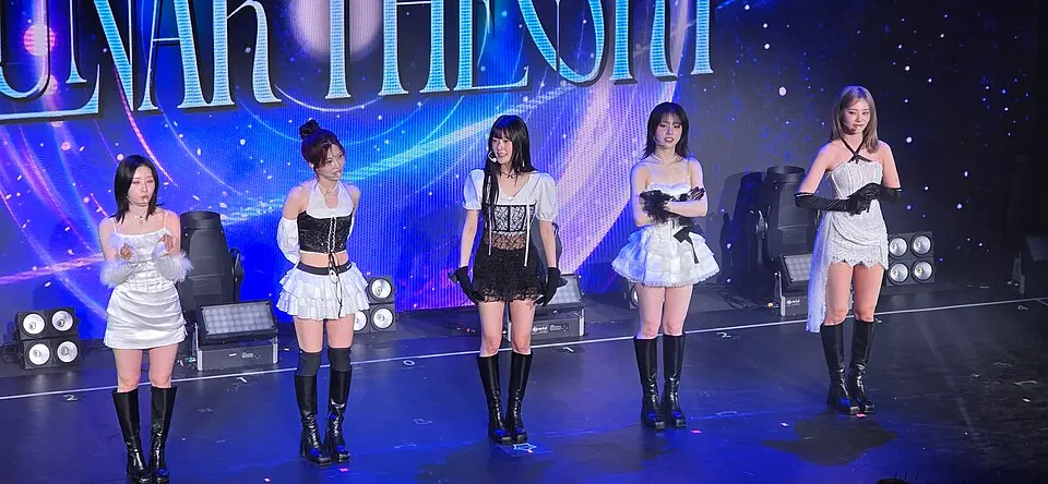 Five young women in black and white stage outfits and boots performing on a stage under bright lights.