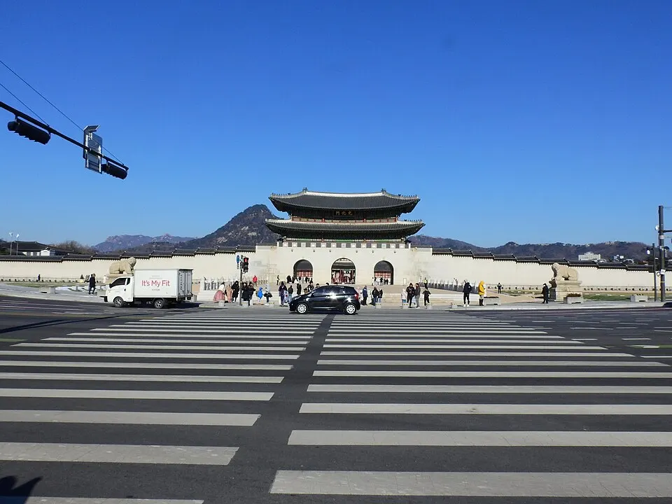A traditional Korean gate with a two-tier tiled roof on a stone base behind a wide pedestrian crossing, with mountains beyond.