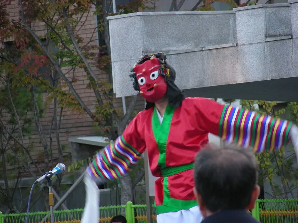 A dancer in a red mask with big white eyes, a red and green costume and long striped sleeves, arms stretched wide outdoors.