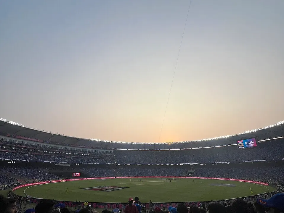A huge cricket stadium at dusk with a glowing sky, packed stands and players far below.