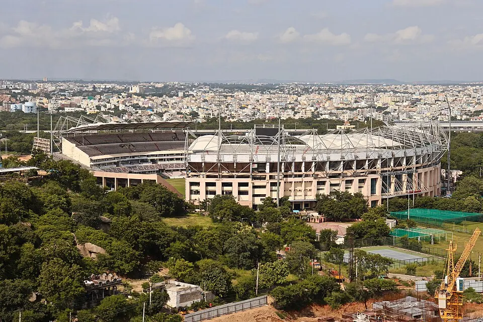 A large cricket stadium with a white roof seen from a hill among trees and city buildings.
