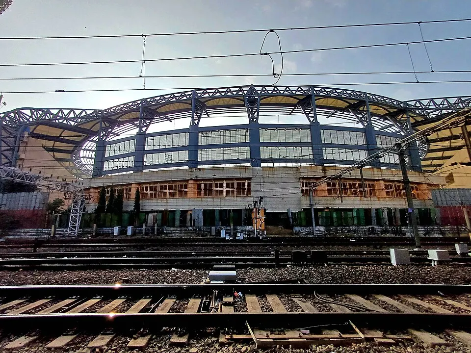 The curved steel roof of a stadium seen from across railway tracks and overhead wires.