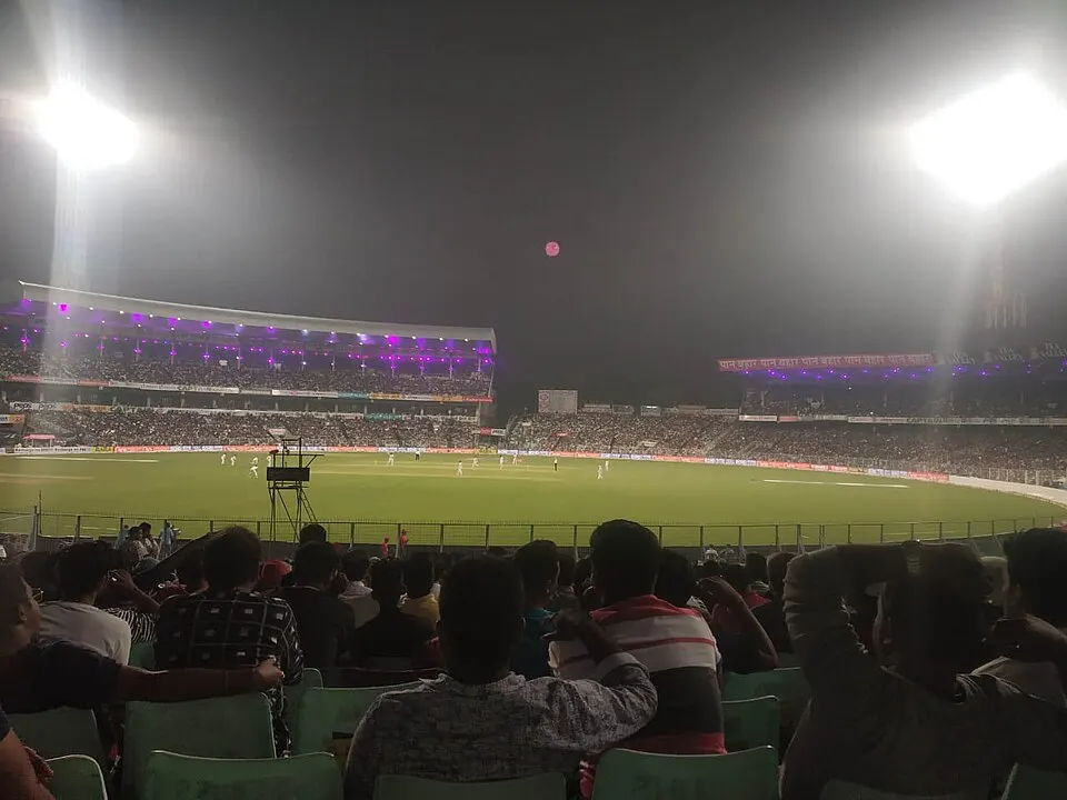 A cricket ground lit at night with floodlights, a crowd in the stands and players in white.