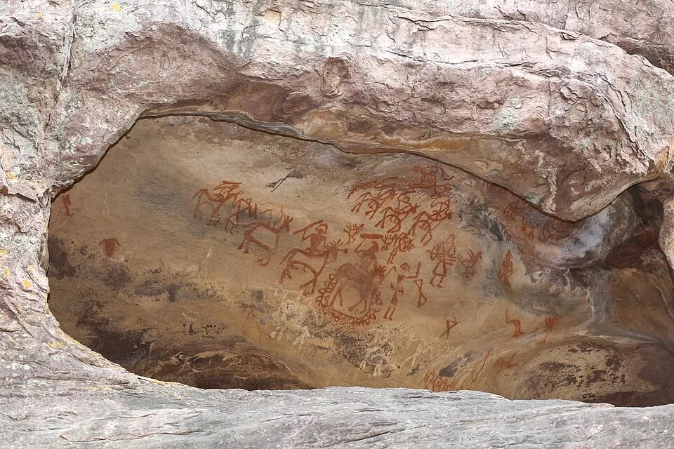 Red-brown paintings of animals and stick-like human figures cover the inside of a rocky overhang.