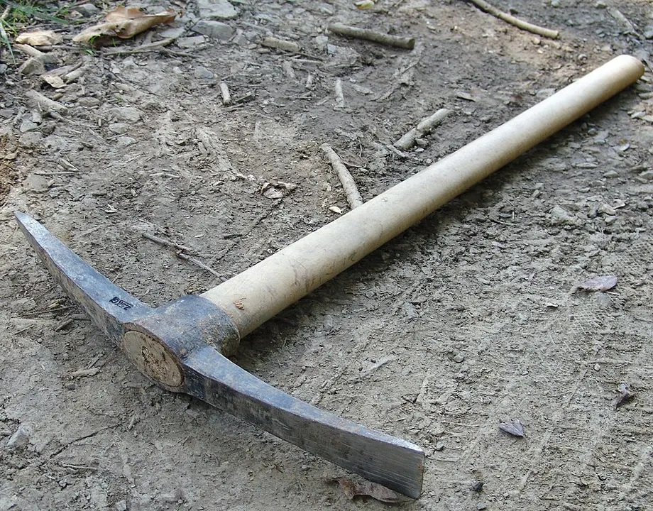 A pickaxe with a long pale wooden handle and an iron head lying on the dusty ground.