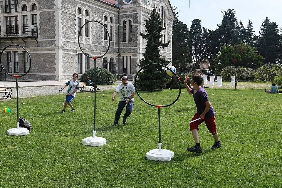 Students playing a ball game on a lawn with tall hoops on poles in front of a large stone building.