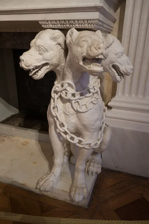 A white marble statue of a chained dog with three snarling heads beside a fireplace.