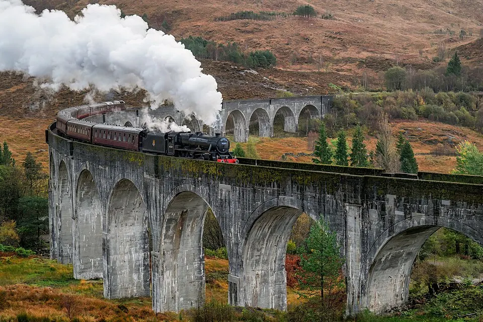 A steam train with a plume of white smoke crossing a long curved stone viaduct among autumn-coloured hills.