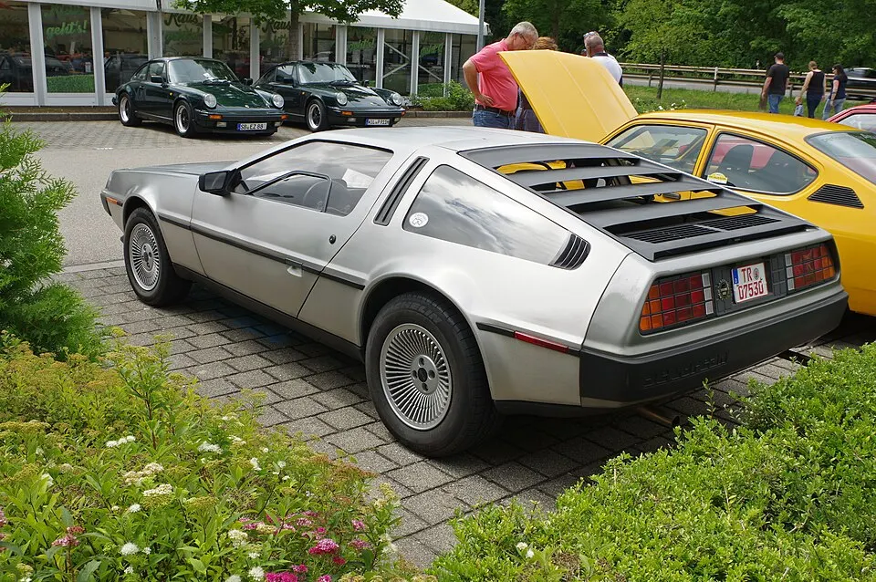 A silver stainless-steel sports car with louvred rear window and wheel covers parked on paving at a car show.