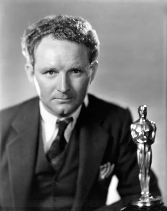 A black-and-white studio portrait of a serious man in a three-piece suit beside a small golden statuette.