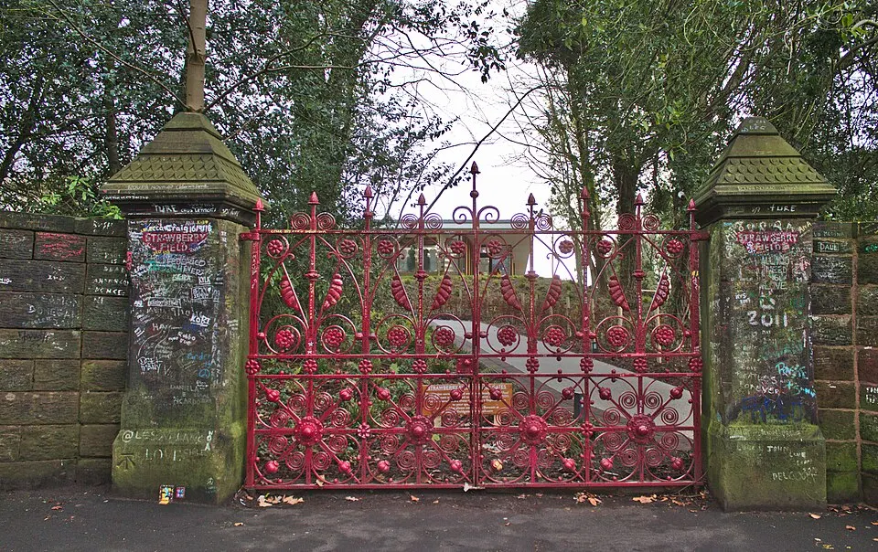 Ornate red iron gates between two weathered stone pillars covered in graffiti, with trees behind them.