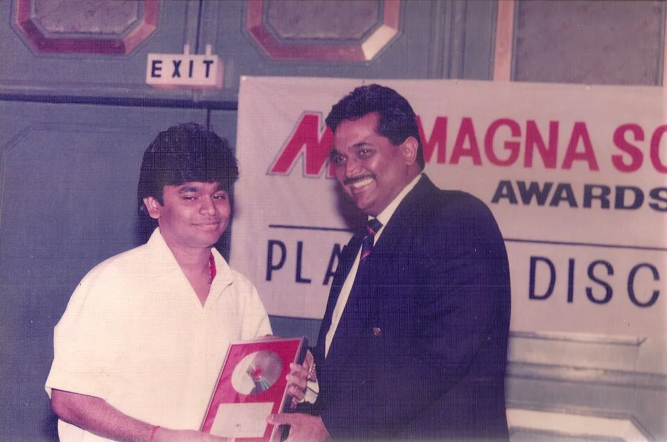 An old colour snapshot of a young man in a white shirt being handed a framed platinum disc by a smiling man in a dark suit at an awards event.