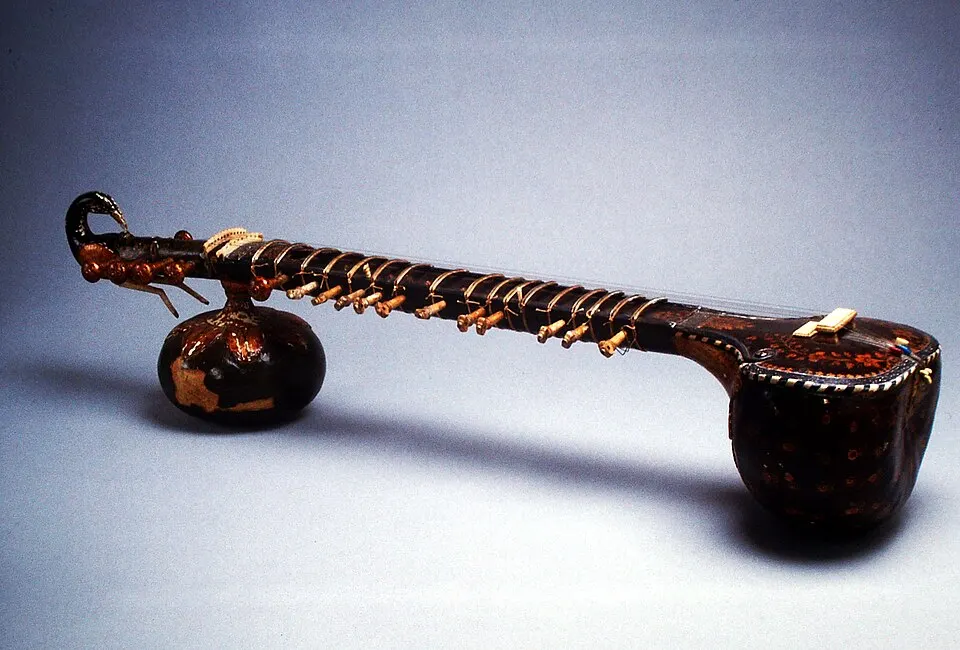 An old Indian string instrument with a long fretted neck, a carved bird-head end and two gourd resonators, on a pale background.