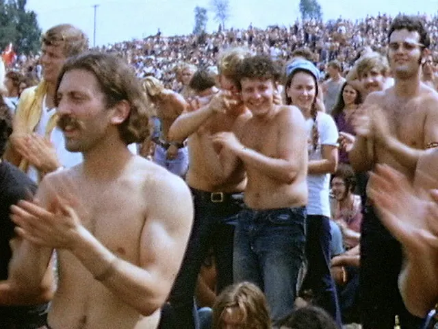 A crowd of young people with long hair, many shirtless, clap their hands on a sunny hillside.