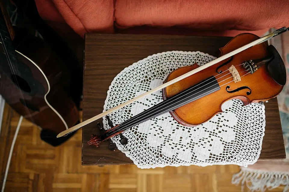 A wooden violin with its bow lies on a white crocheted doily on a small table, seen from above.
