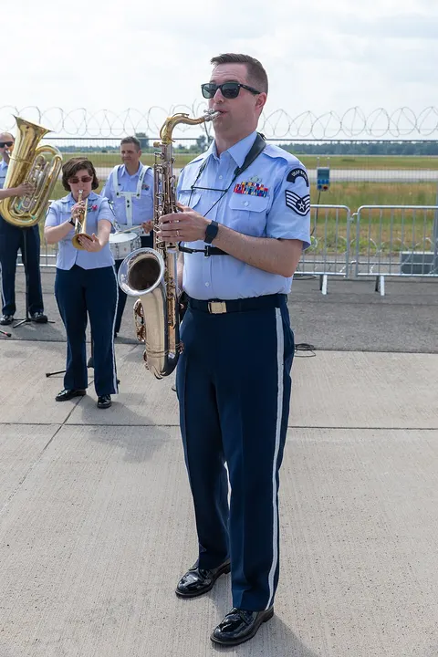 A uniformed band member with sunglasses plays a golden saxophone outdoors, with a trumpeter and drummer behind him.