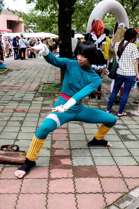 A cosplayer in a teal bodysuit with yellow leg warmers and bandaged hands in a wide fighting stance on a paved square.