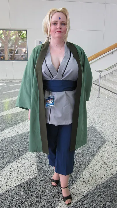 A cosplayer with long blond hair, a blue diamond mark on the forehead and a green open coat over a grey top and blue sash.