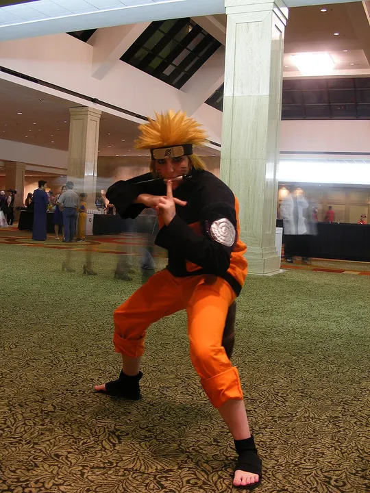 A cosplayer in an orange and black tracksuit with a spiky blond wig and a metal forehead headband, crouching and gripping a kunai in a hall.