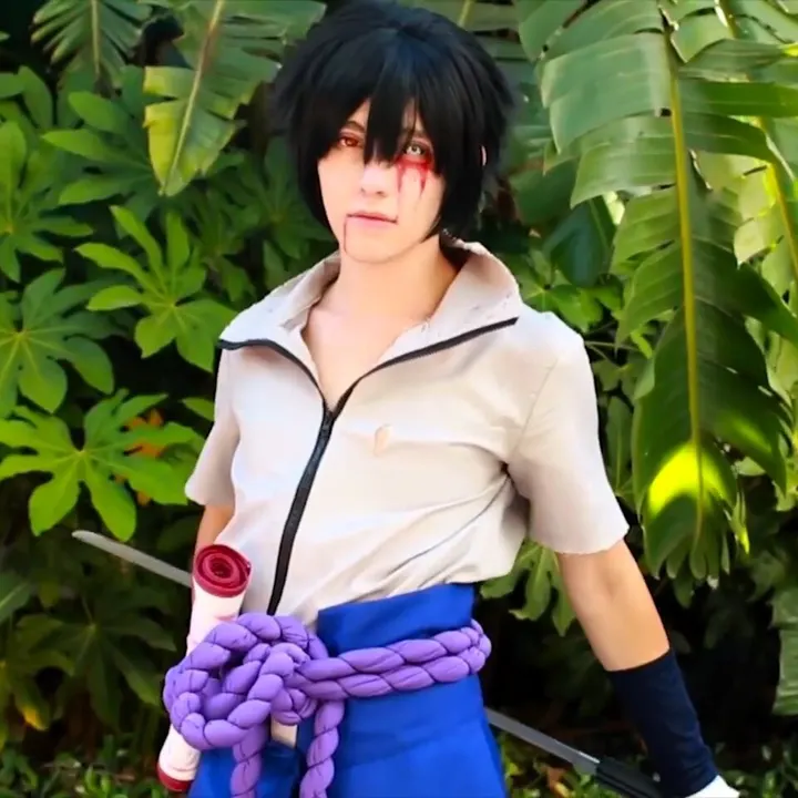 A cosplayer with black hair and a red-marked eye, in a cream high-collared top with a thick purple rope belt and a sword, in front of green leaves.
