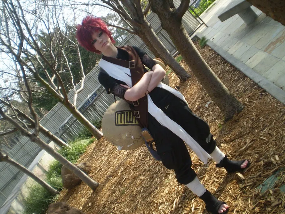A cosplayer with short red hair and folded arms, wearing a black outfit with a cream sash and a large gourd on the back, on a wood-chip path.