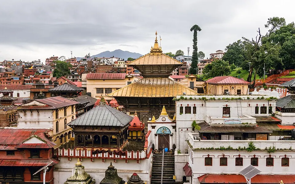 A golden-roofed pagoda-style temple among white and red buildings, with a city and hills behind.