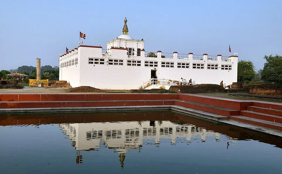 A white temple with prayer flags beside a still pond that reflects it, with a stone pillar to one side.