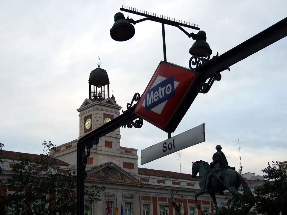 A clock tower on a pale building behind a red Metro sign and a bronze statue of a horseman.