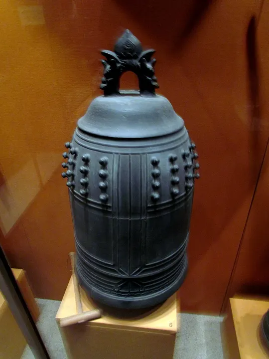 A dark bronze temple bell with rows of knobs on its sides and a wooden striker in front, in a museum case.