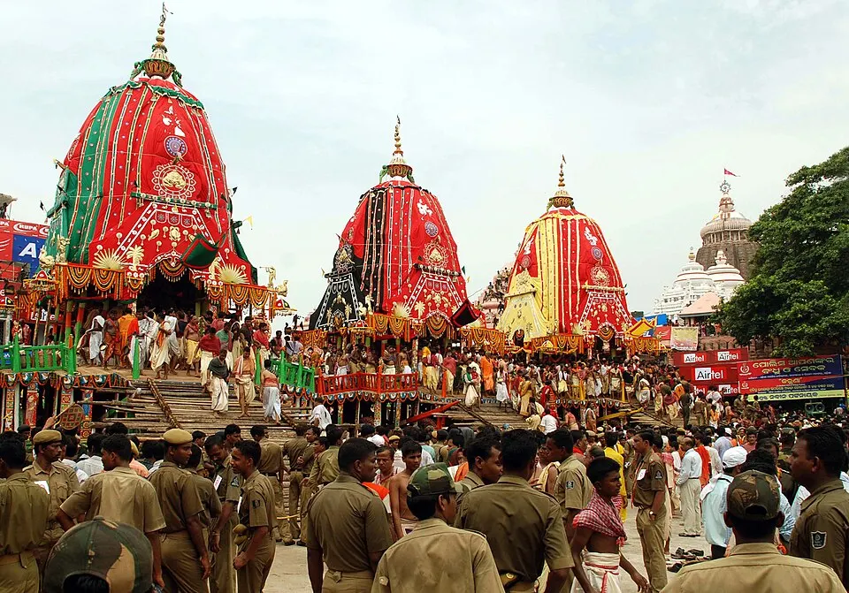 Three huge chariots with red, green and yellow canopies stand in a crowd of devotees and uniformed police outside a white temple.