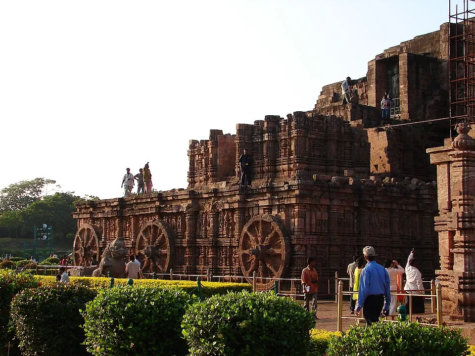 A ruined sandstone temple shaped like a chariot with large carved stone wheels along its base, with visitors walking around it.