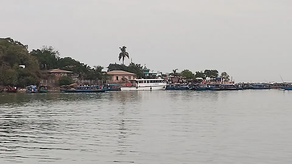 A white passenger boat and many small boats are moored along a green island shore on a calm, wide lake.